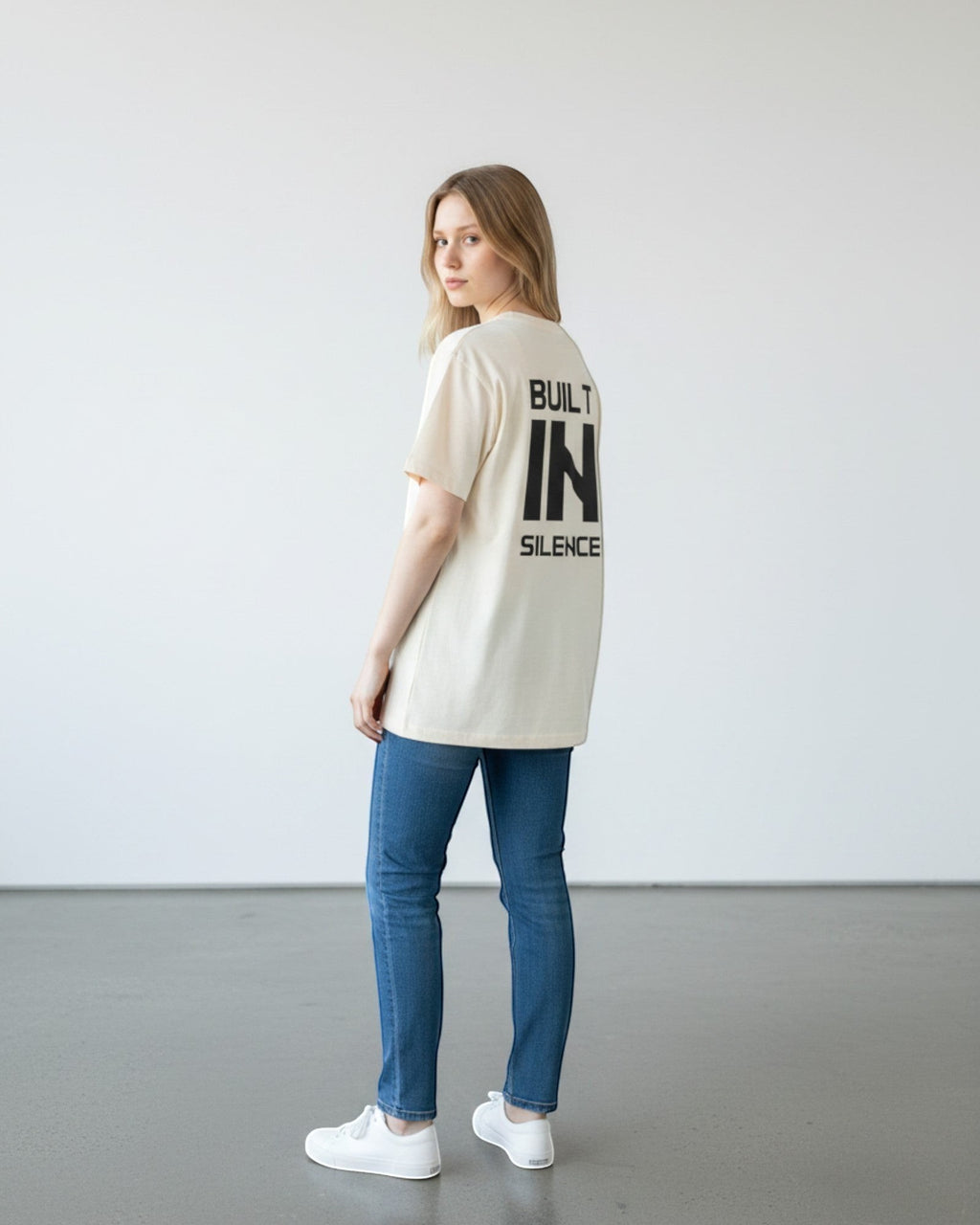FALLR wear cream 100% cotton oversized t-shirt with black "BUILT IN SILENCE" graphic print on back, worn by a woman with blue jeans.