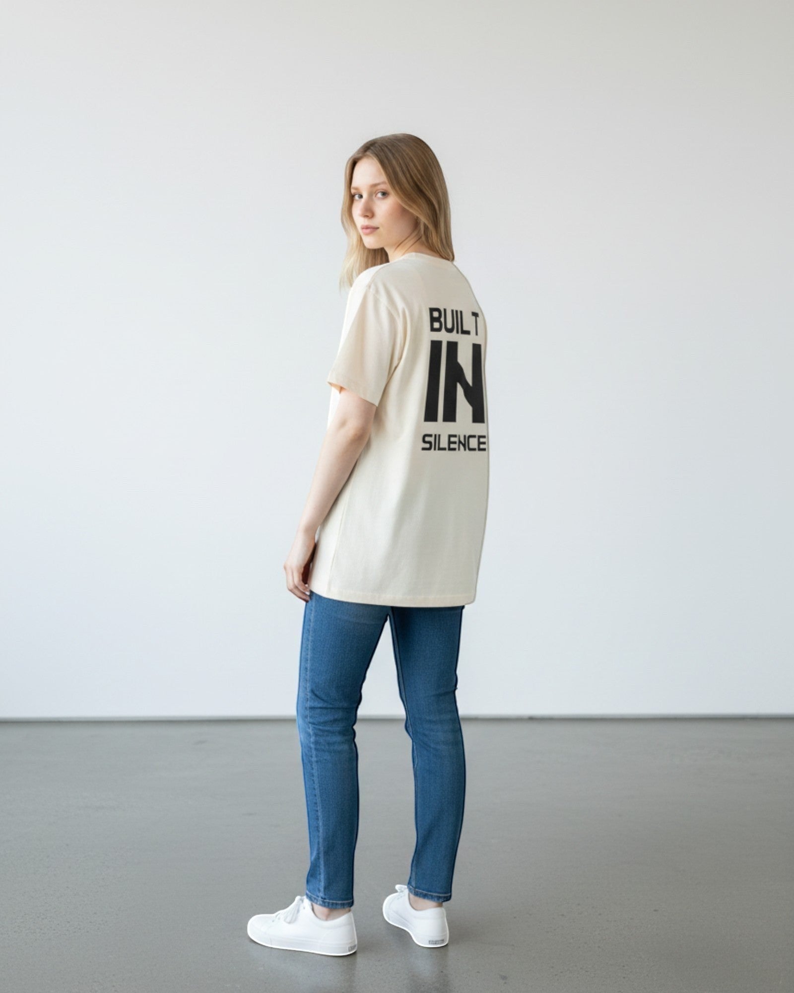 FALLR wear cream 100% cotton oversized t-shirt with black "BUILT IN SILENCE" graphic print on back, worn by a woman with blue jeans.
