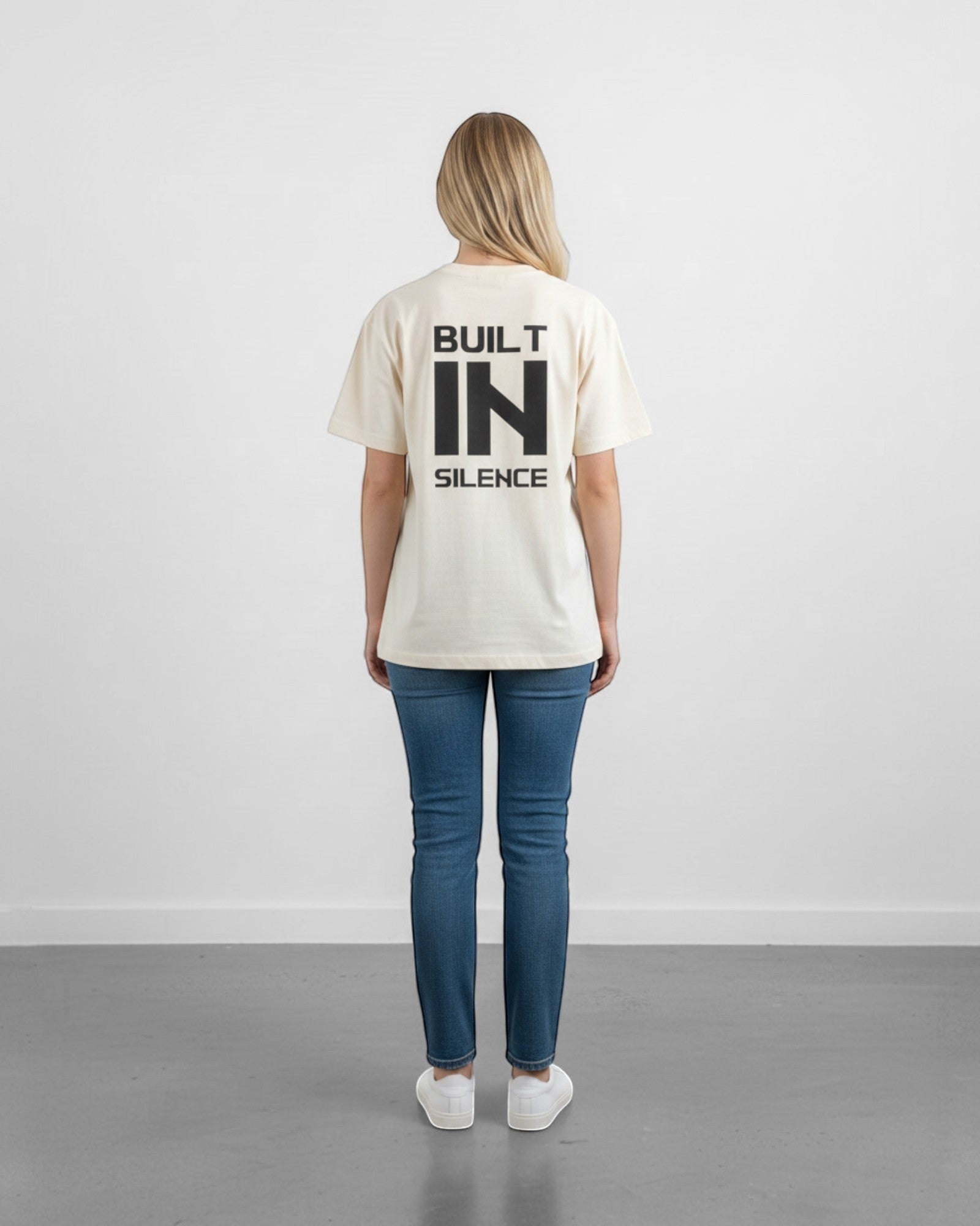 FALLR wear cream 100% cotton oversized t-shirt with black "BUILT IN SILENCE" graphic print on back. Model wears jeans and white sneakers.